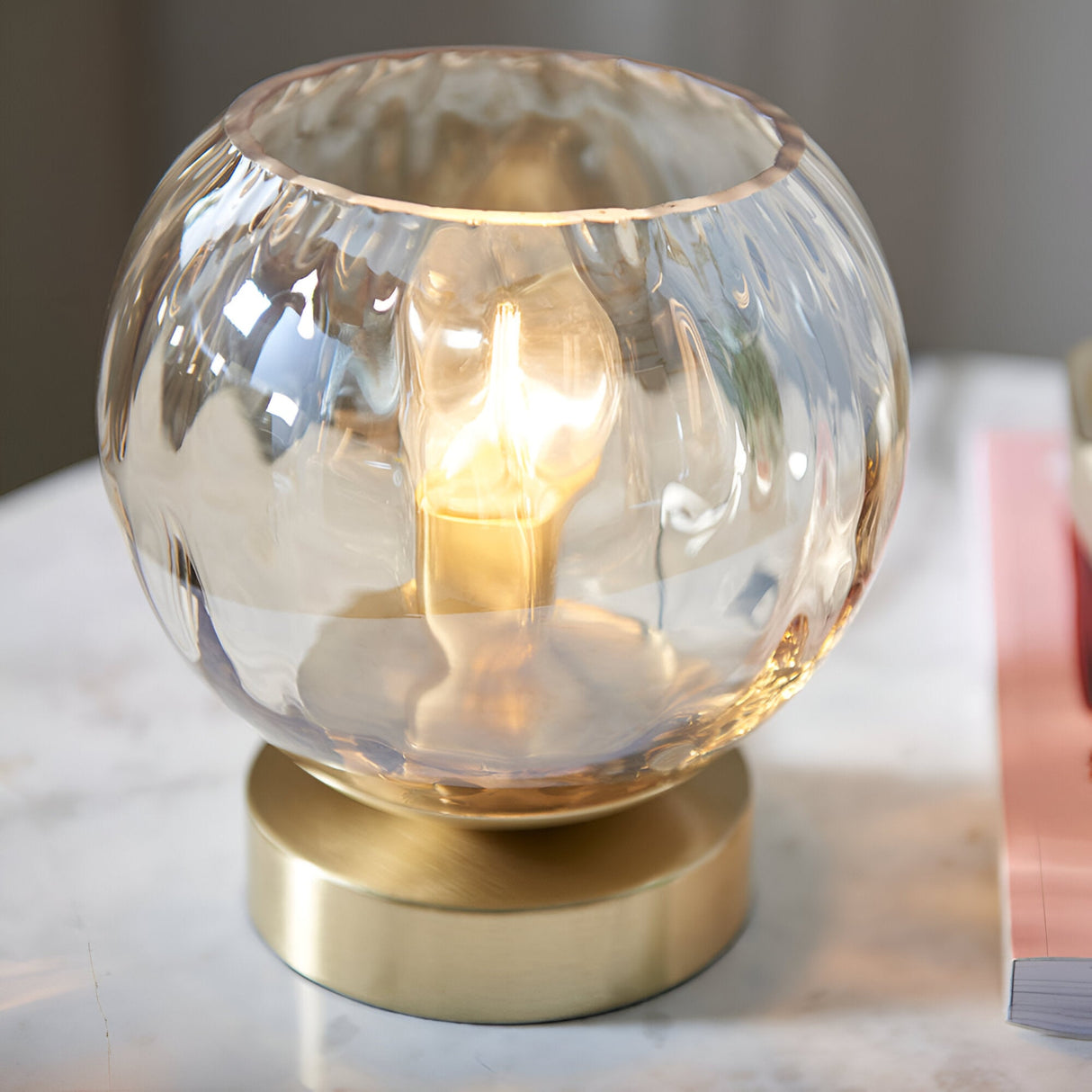 Draycott Dimpled Glass Table Lamp in Satin Gold, with a champagne-toned dimpled glass globe shade on a satin gold base, lit and shown on a marble surface.