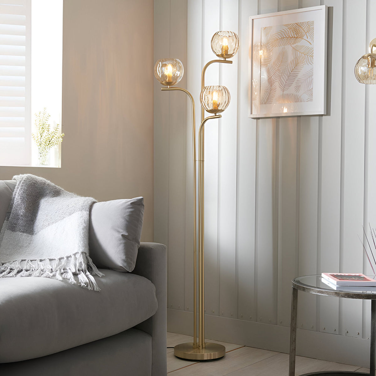 Endon Draycott 3 light floor lamp with champagne dimpled glass globe shades on a satin brass stem, styled beside a grey sofa in a living room corner.