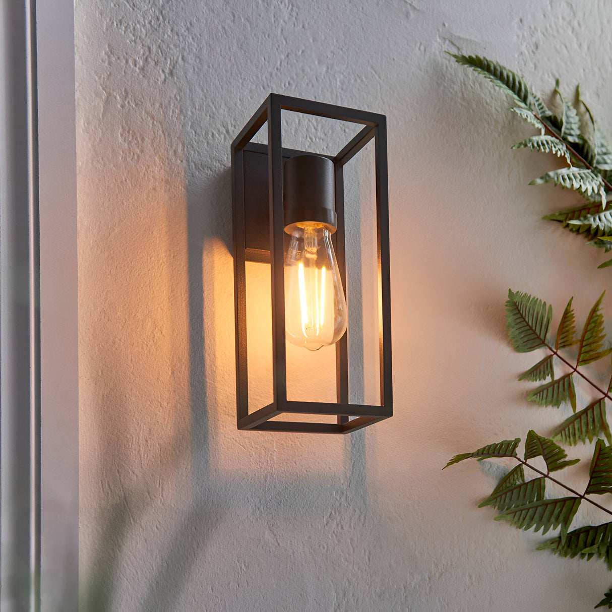 Brenford Outdoor Wall Light in black, an open rectangular aluminium cage design with visible E27 filament bulb, mounted on a textured wall near foliage.