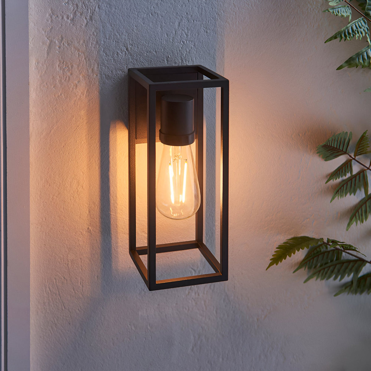 Brenford Outdoor Wall Light in black aluminium with an open lantern-style frame, E27 filament bulb visible, mounted on a textured wall near foliage.