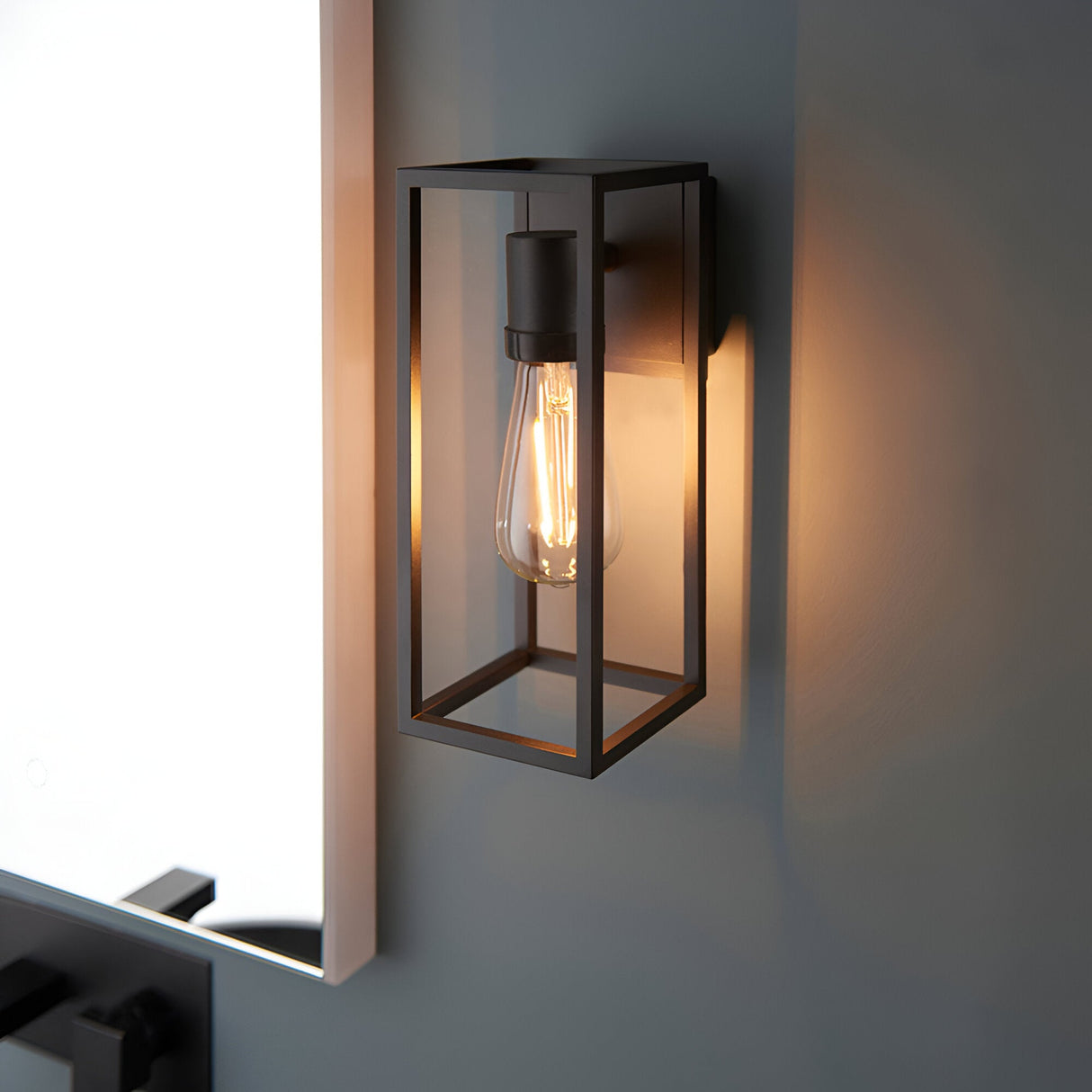 Brenford Outdoor Wall Light in black aluminium with an open lantern-style frame and exposed filament bulb, mounted on a grey wall.