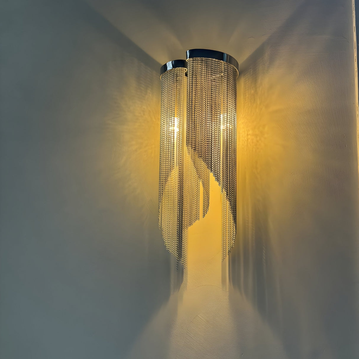 The Valora 2 Light Metal Chain Veil Wall Light in Gold features cascading metallic chains that emit warm light, creating intricate reflections and shadows on a smooth, light-colored wall.