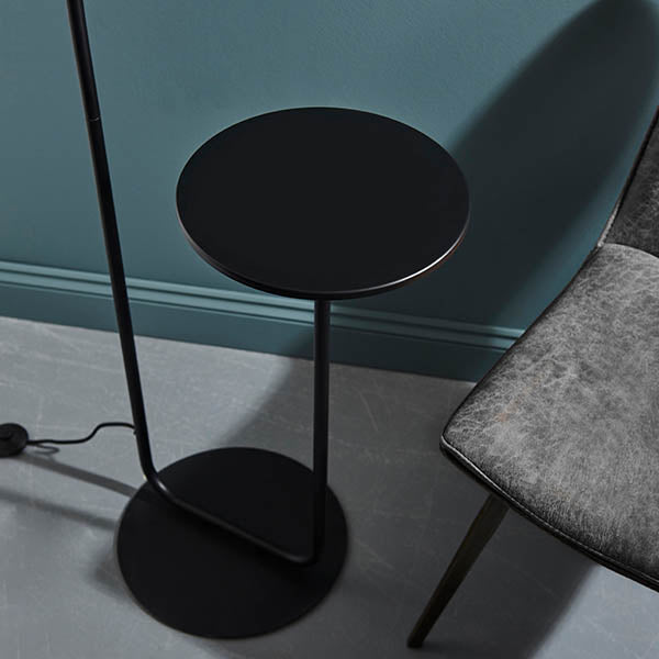 Caldfield Floor Lamp in Black with round metal base and slim stem, featuring a built-in round black table top, shown beside a chair.