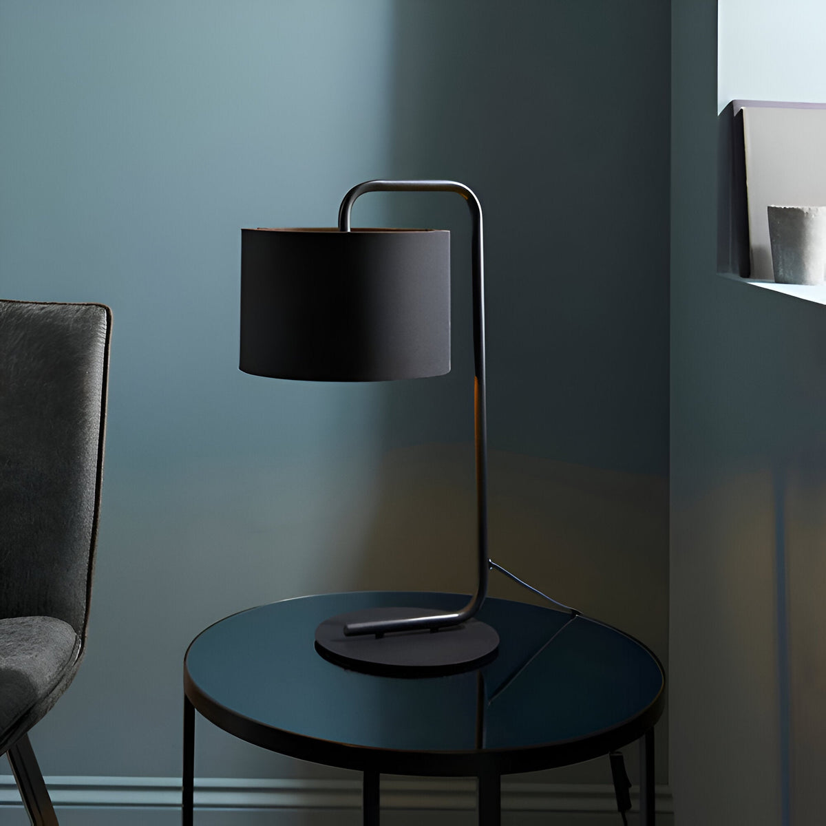 Caldfield Table Lamp in satin black with a curved metal stem and cylindrical fabric shade, shown on a dark round side table in a muted living room setting.