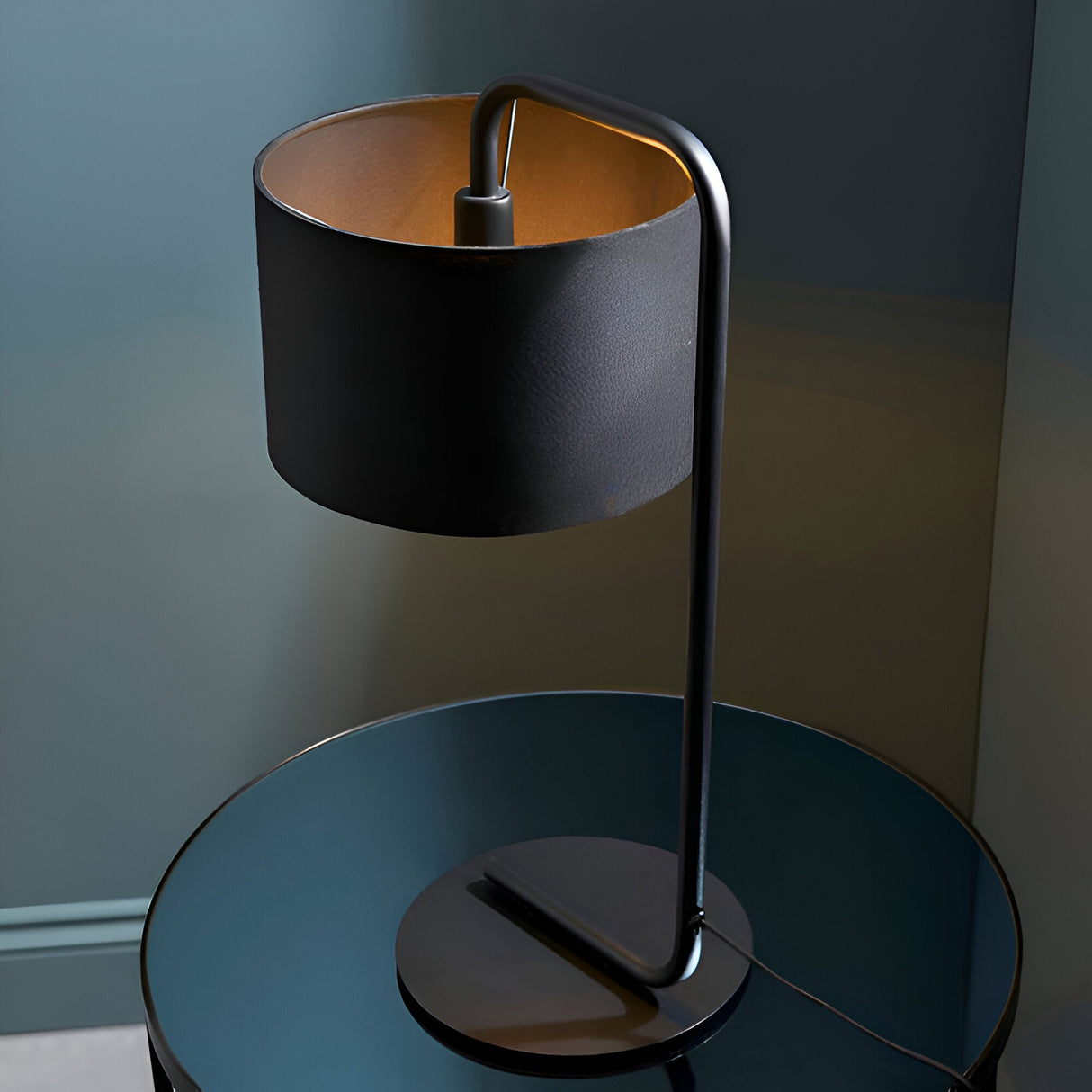 Caldfield Table Lamp in satin black metal with a fabric drum shade, shown on a glass side table against a dark background.