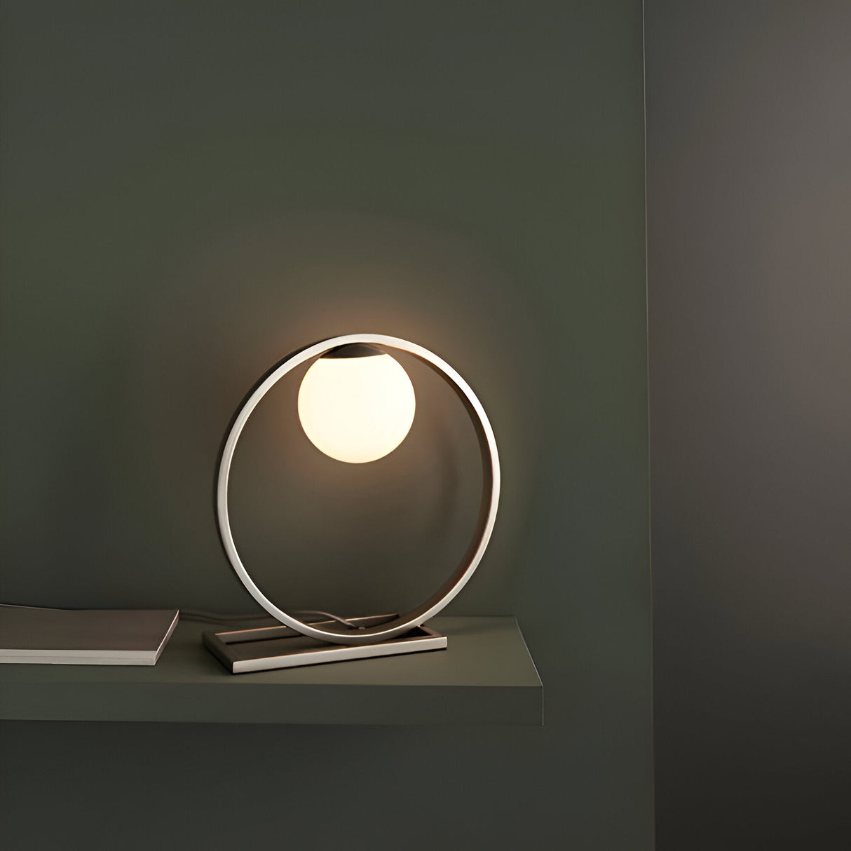 Brackstead Table Lamp in brushed silver, a circular metal frame on a rectangular base holding a glowing opal glass sphere, shown lit on a shelf.