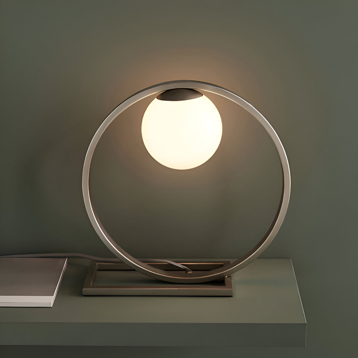 Brackstead Table Lamp in brushed silver, a circular metal ring on a rectangular base holding a glowing opal glass sphere, shown on a shelf.