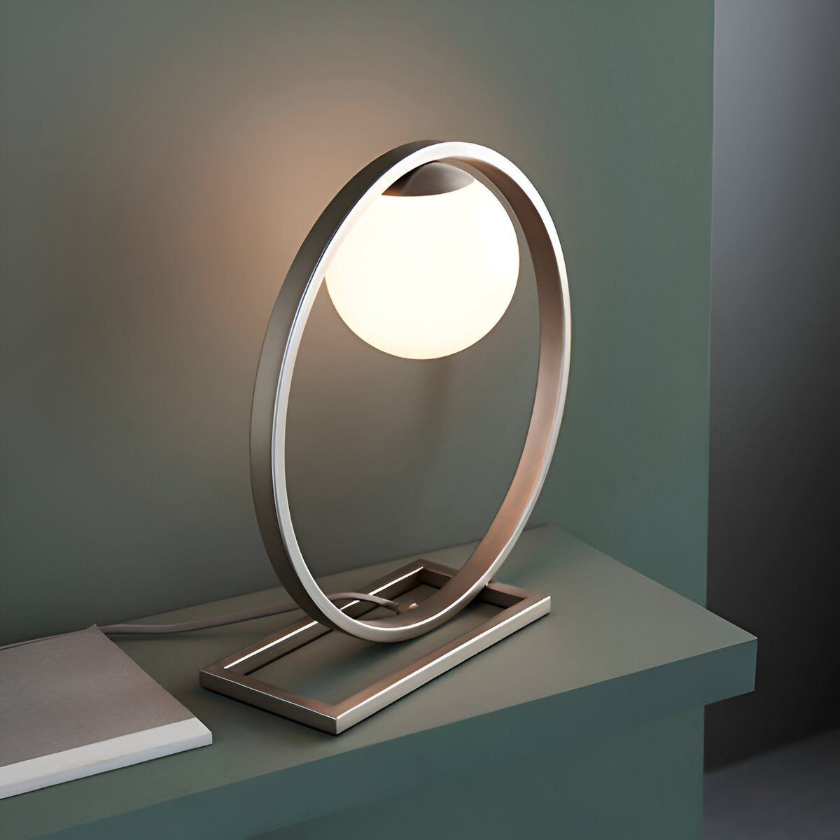 Brackstead Table Lamp in brushed silver, a circular frame on a rectangular base holding a glowing opal glass sphere, shown lit on a shelf.