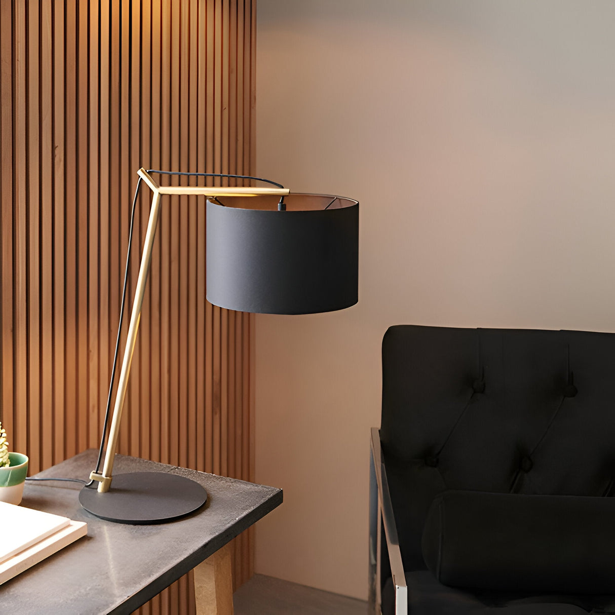 Endon Carlfield cantilevered table lamp with matt brass arm, black cotton drum shade and matt black base, on a desk beside a dark armchair.