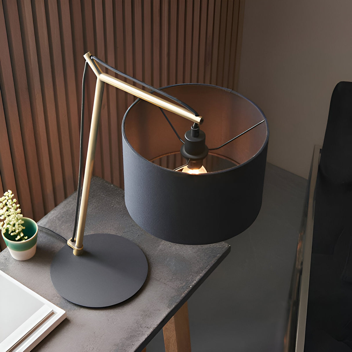 Endon Carlfield cantilevered table lamp with matt brass arm, black cotton drum shade and matt black base on a dark desk beside a small potted plant.