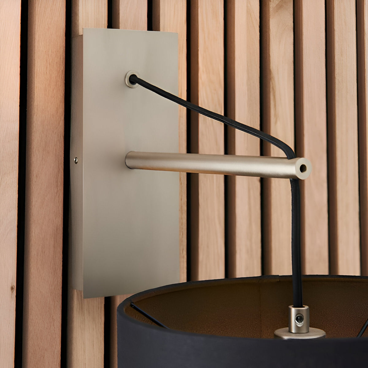 Carlfield Wall Light in Nickel, showing the matt nickel rectangular backplate, cantilevered stem, black fabric cord and shade, against a wooden slatted wall.