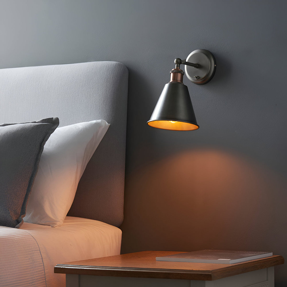 Endon Brackley wall light in aged copper and pewter with an adjustable conical shade, mounted above a wooden bedside table beside a grey headboard.