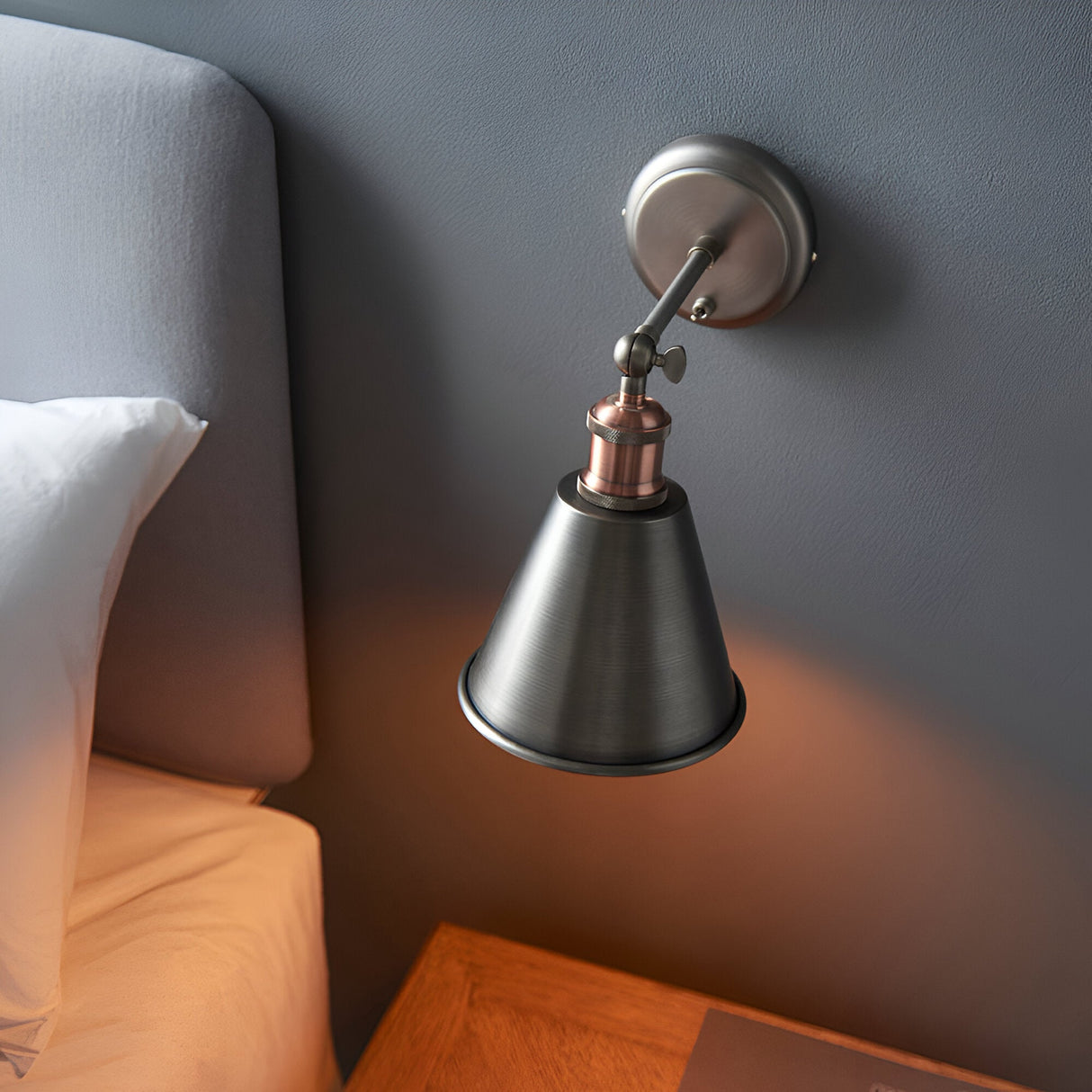 Endon Brackley wall light with adjustable conical shade in aged pewter and copper, mounted on grey bedroom wall beside upholstered headboard.