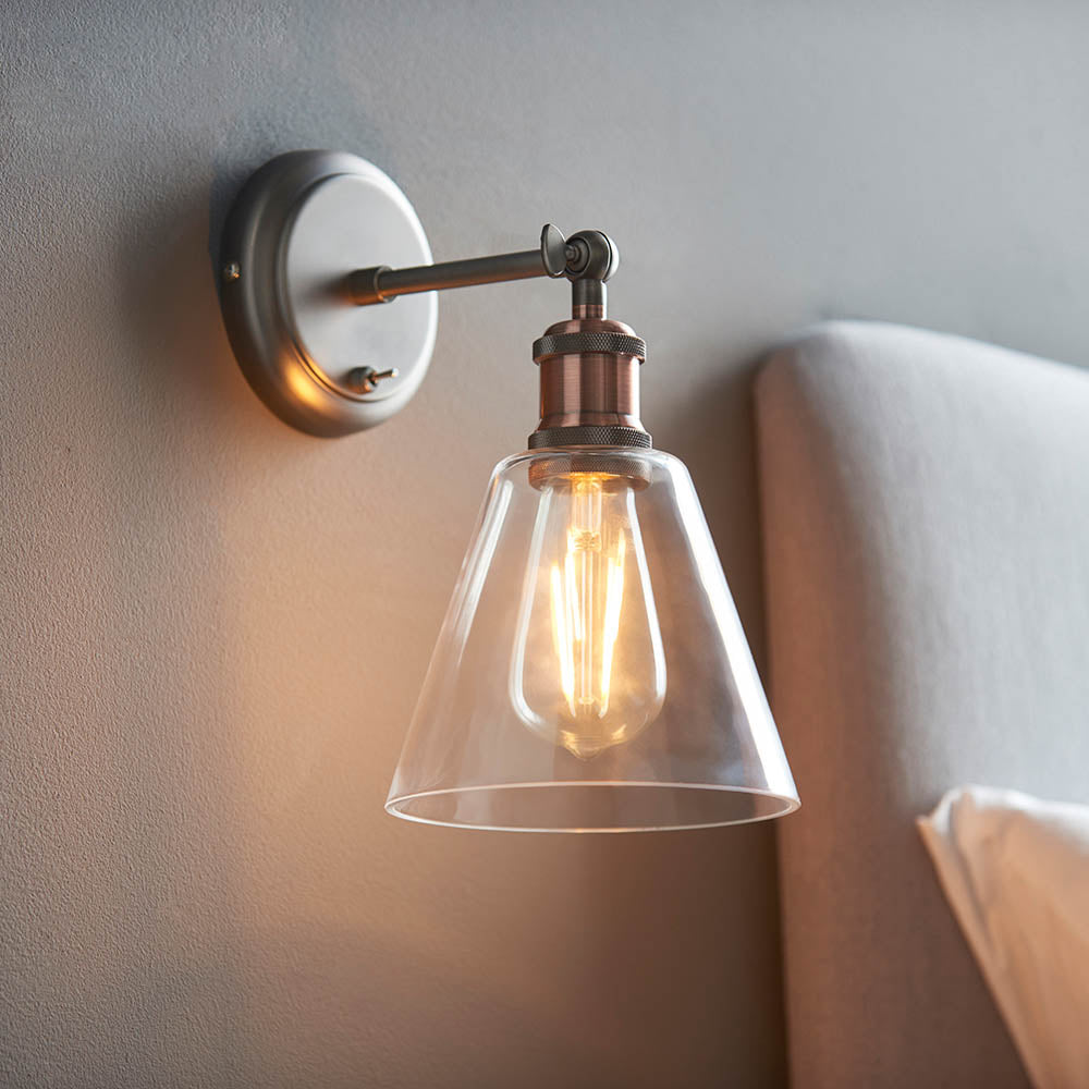 Brackley glass wall light in pewter and copper finish with cone-shaped clear glass shade and exposed bulb, mounted above a bed headboard.