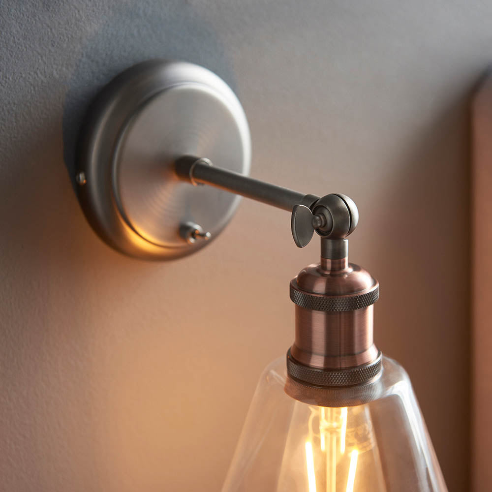 Brackley glass wall light in aged pewter and copper, with round wall plate, adjustable arm, and clear glass shade housing an exposed bulb.