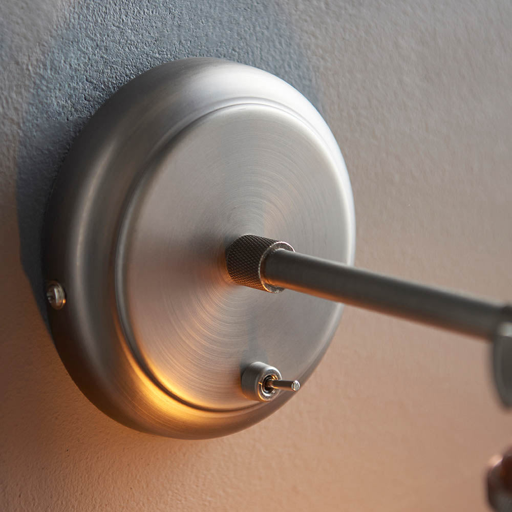 Close-up of the Brackley glass wall light's circular pewter backplate with knurled adjustable joint and extending arm, mounted on a textured wall.