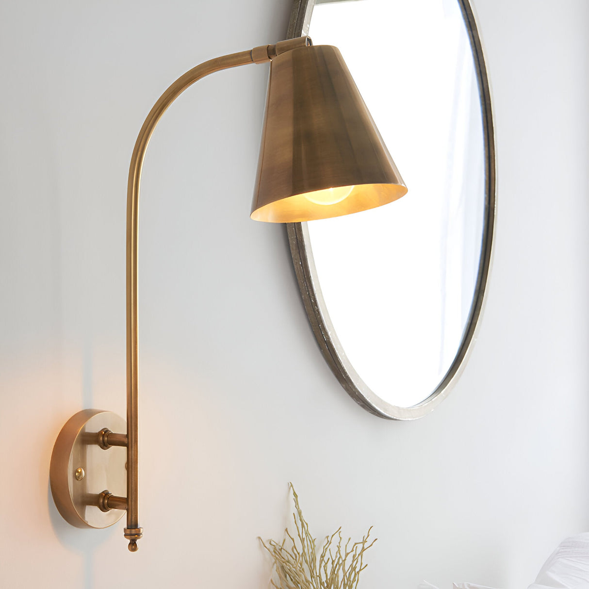 Chartmere Wall Light in antique brass with tall curved arm and angled cone shade, mounted beside an oval mirror on a white wall.