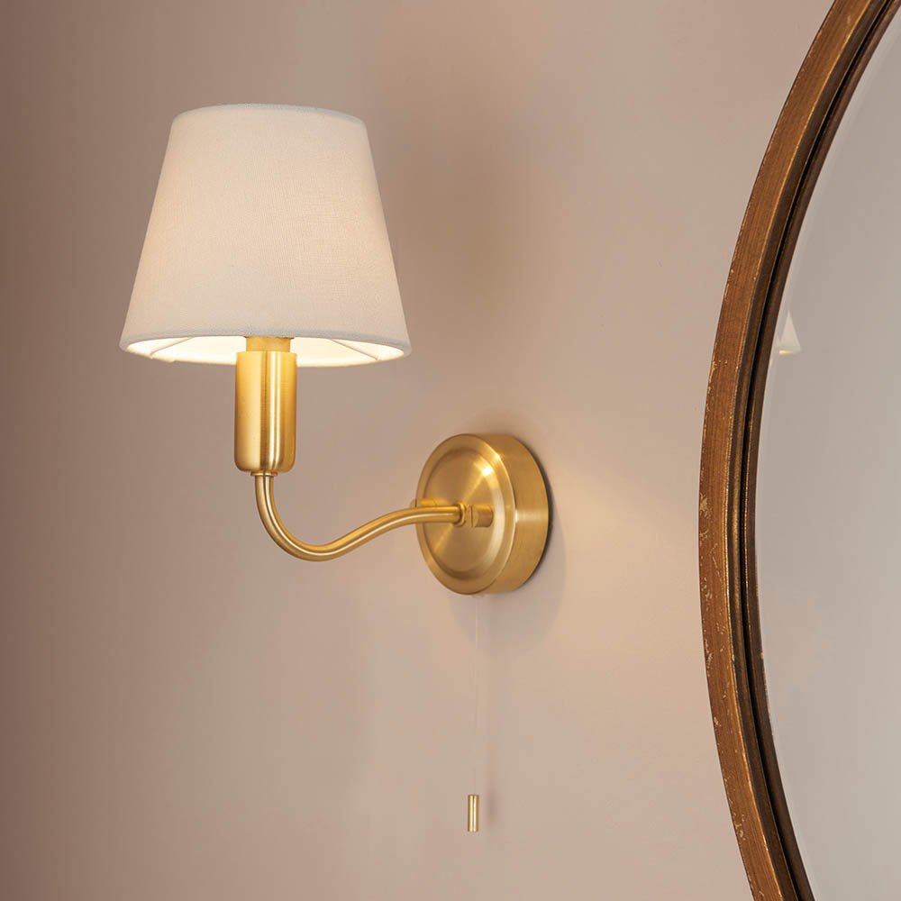 Endon Bevton bathroom wall light in satin brass with a curved arm, ivory fabric shade and pull cord, mounted beside a round brass-framed mirror.