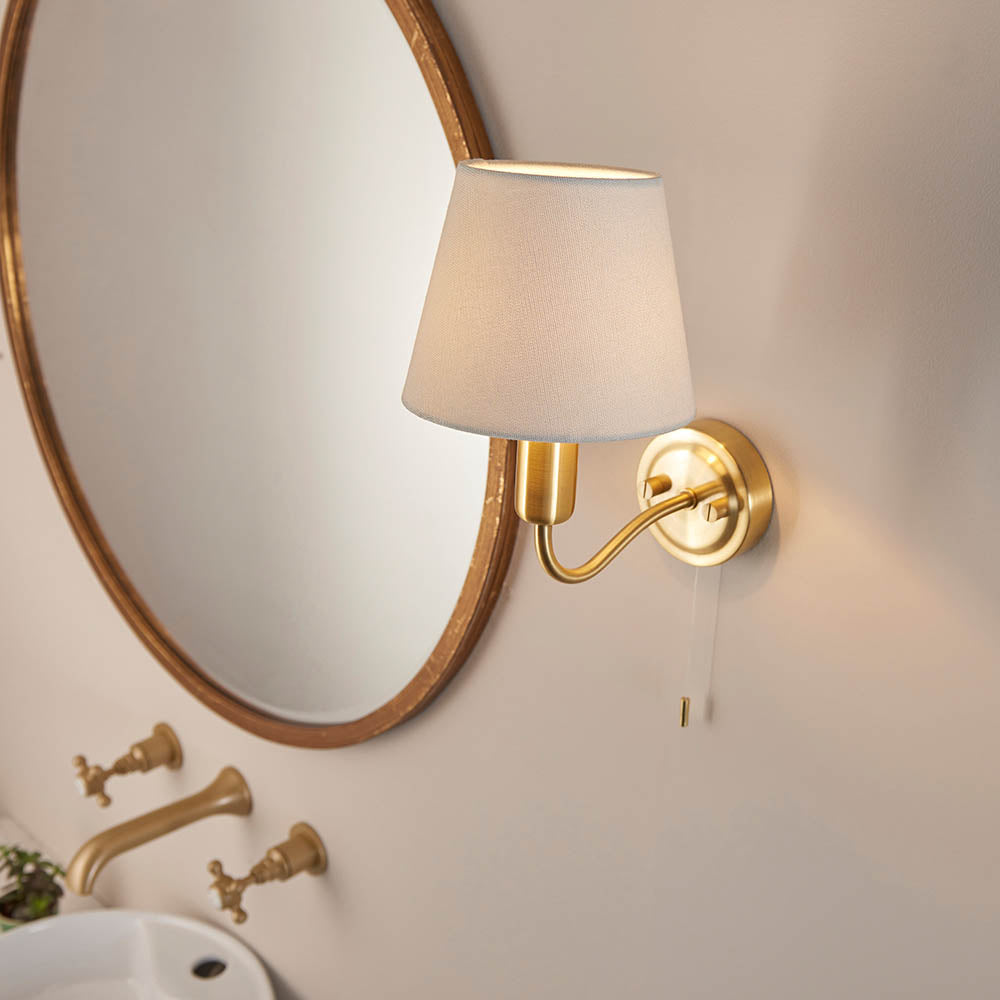 Endon Bevton bathroom wall light in satin brass with a curved arm, ivory fabric shade and pull cord, mounted beside an oval brass mirror above a basin.