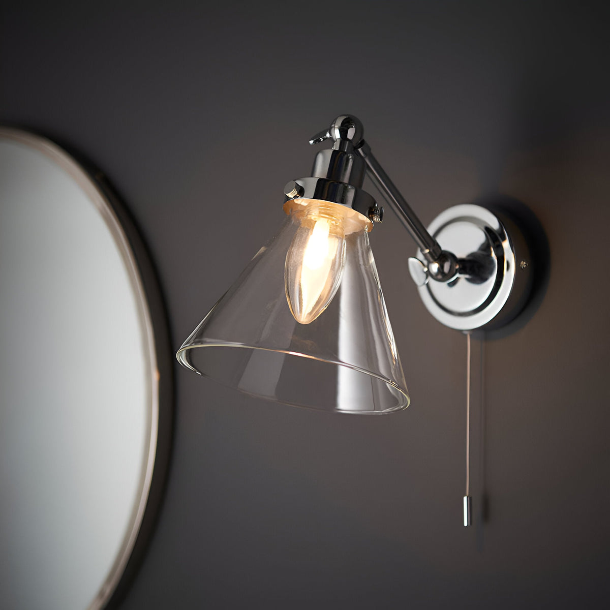 Endon Brackwood bathroom wall light in chrome with clear glass conical shade, adjustable head and pull cord switch, mounted beside a round mirror.