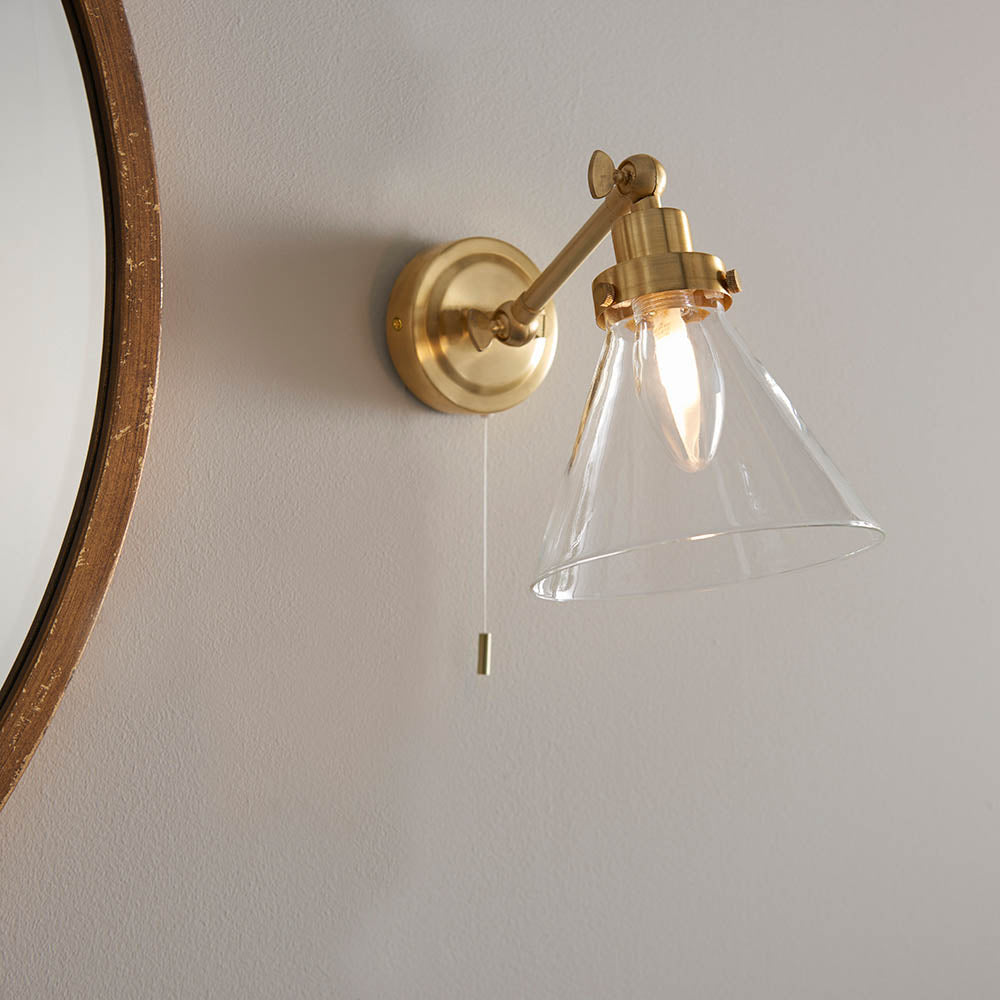 Endon Brackwood bathroom wall light in brass with adjustable arm, clear glass cone shade, G9 bulb and pull cord, mounted beside a round wooden mirror.