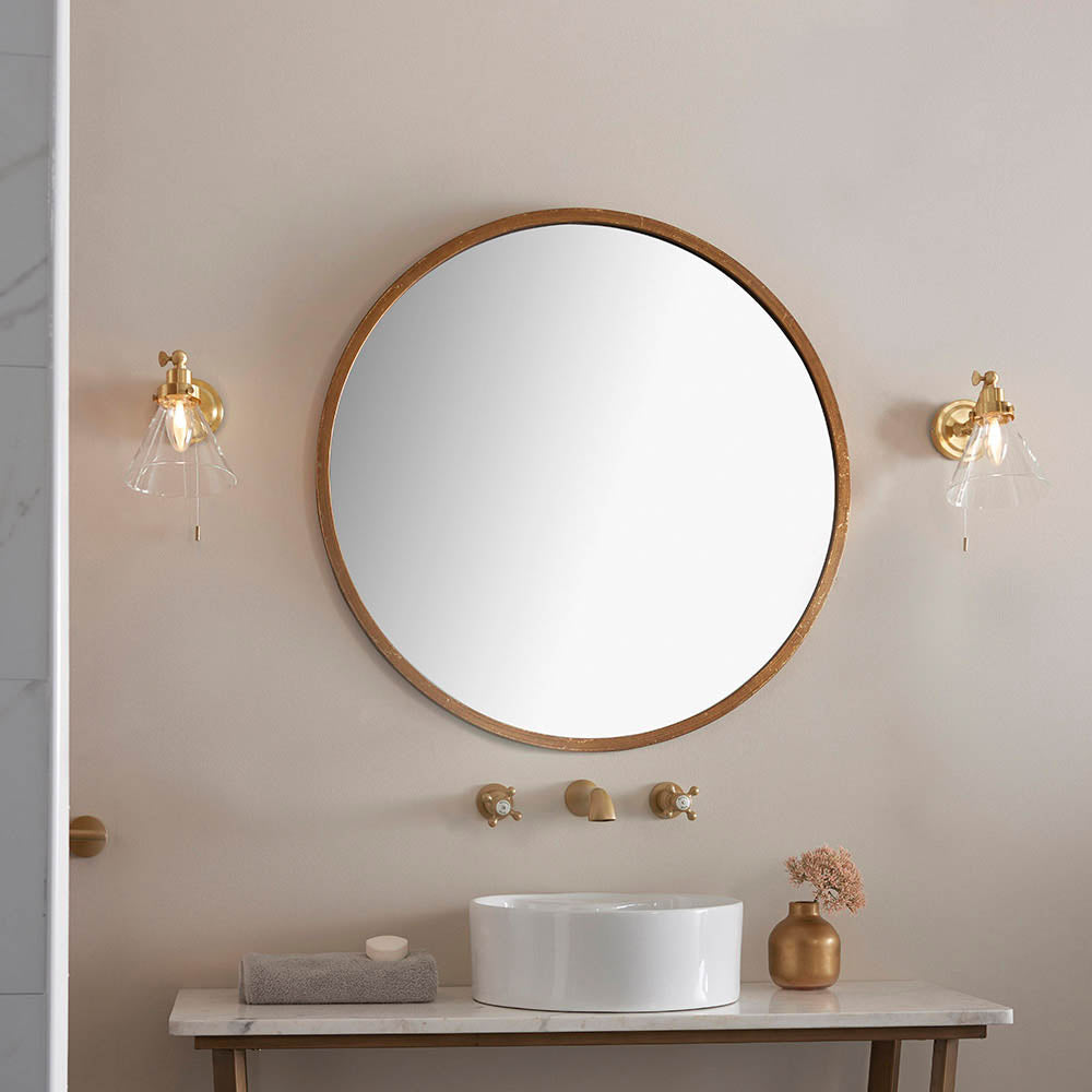 Endon Brackwood bathroom wall lights in brass with clear tapered glass shades, mounted either side of a round wood-framed mirror above a basin.