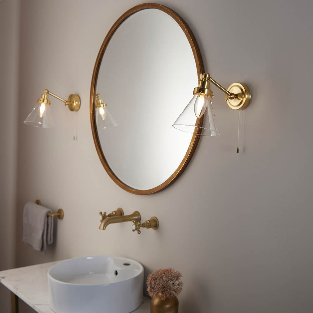 Endon Brackwood bathroom wall lights in brass with clear conical glass shades and pull cords, mounted either side of an oval brass mirror above a basin.