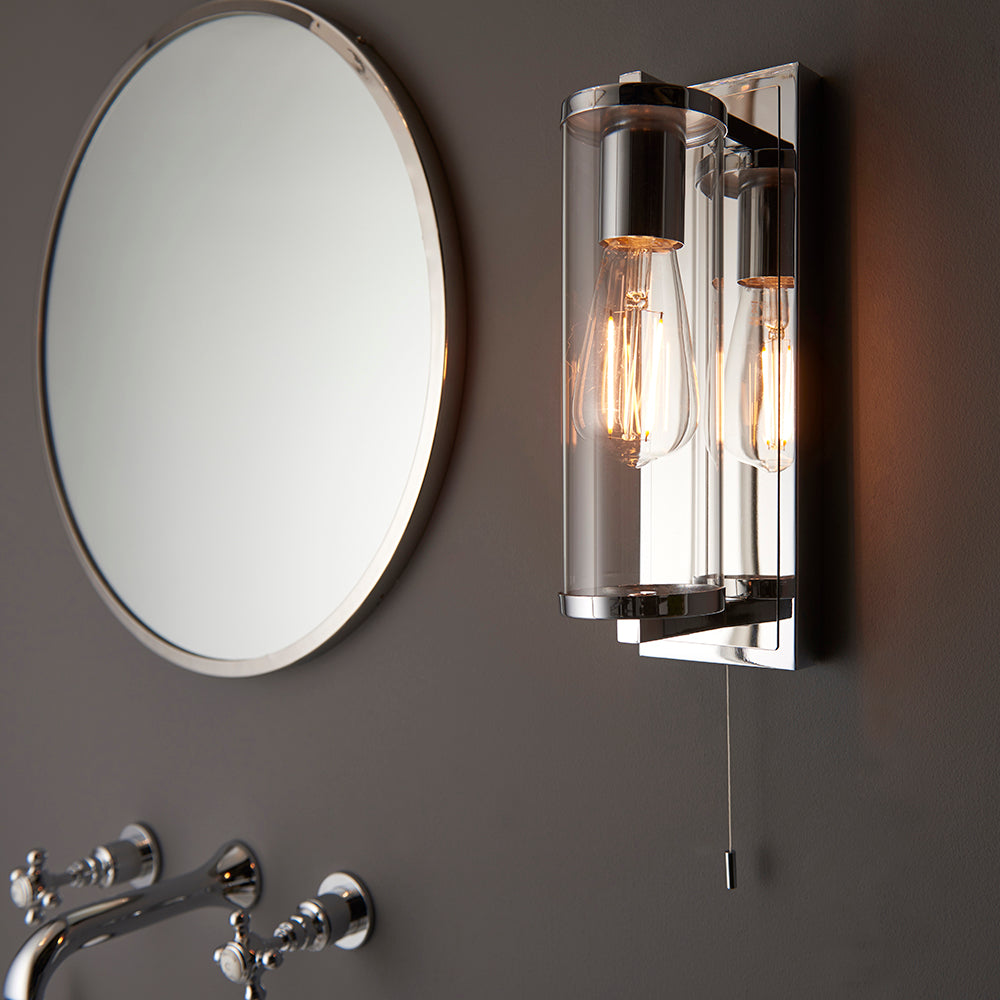 Endon Cranfield chrome bathroom wall light with clear glass tube shade and pull cord, mounted beside a round mirror and chrome basin tap on a grey wall.