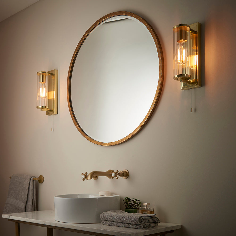 Endon Cranfield bathroom wall lights in brass with ribbed clear glass tube shades and pull cords, mounted either side of a round mirror above a basin.
