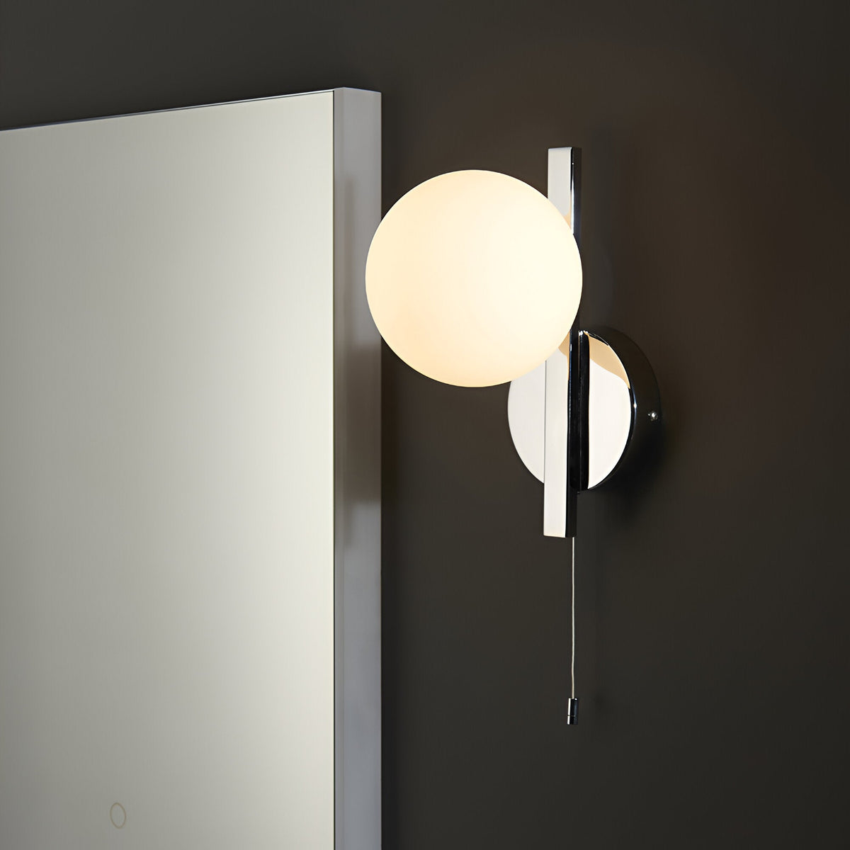 Endon Kelham bathroom wall light with a matt opal glass sphere on a chrome geometric frame with pull cord, mounted beside a mirror on a dark wall.