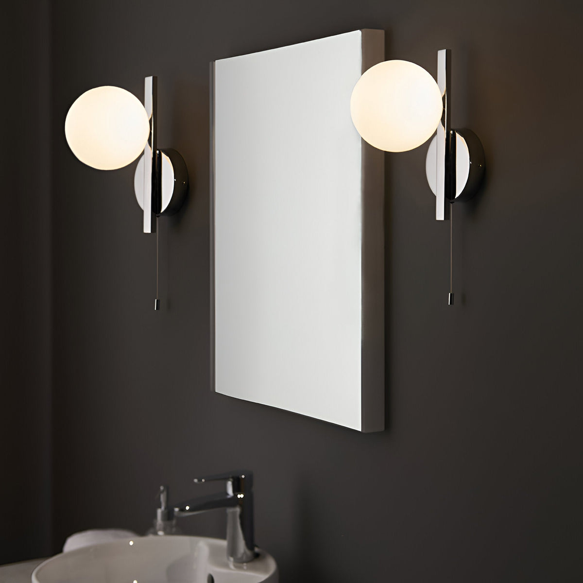 Endon Kelham chrome bathroom wall lights with opal glass globes and pull cords, mounted either side of a rectangular mirror above a basin on a dark wall.