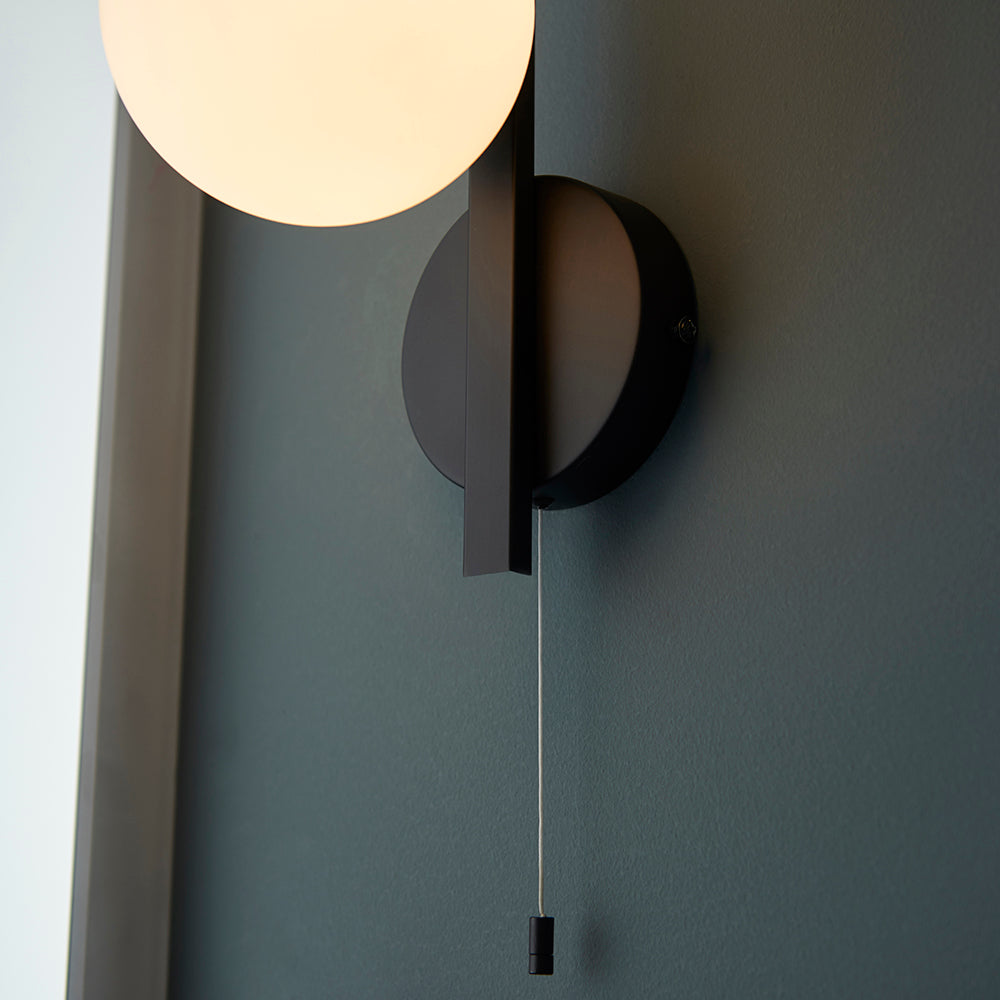 Kelham Bathroom Wall Light in black with opal glass globe shade, geometric metal backplate and pull cord switch, mounted on a dark grey wall.