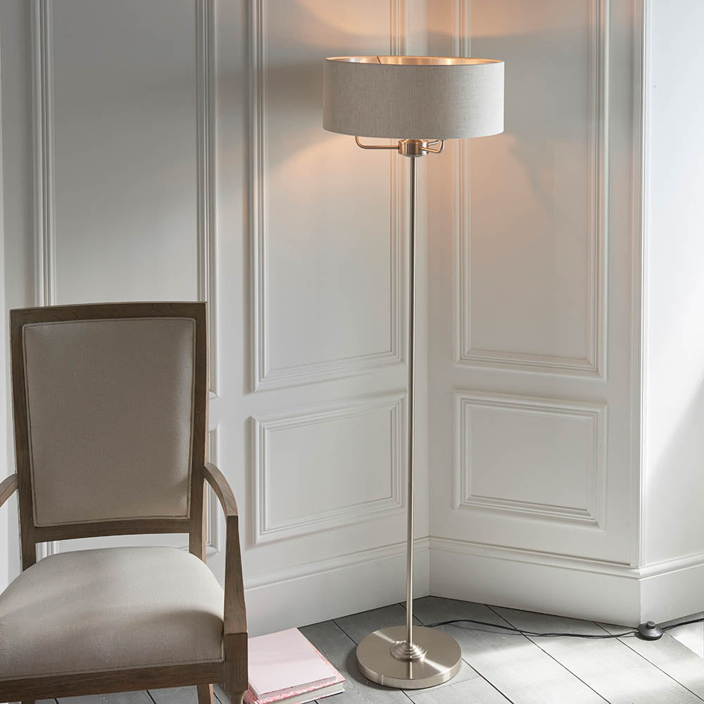 Brenley 3 Light Floor Lamp in brushed chrome with a wide natural linen drum shade, shown beside an upholstered armchair in a living room corner.