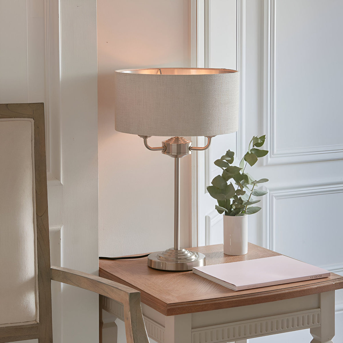 Endon Brenley 3 Light Table Lamp in brushed chrome with natural linen drum shade, styled on a wooden console with eucalyptus and notebook.