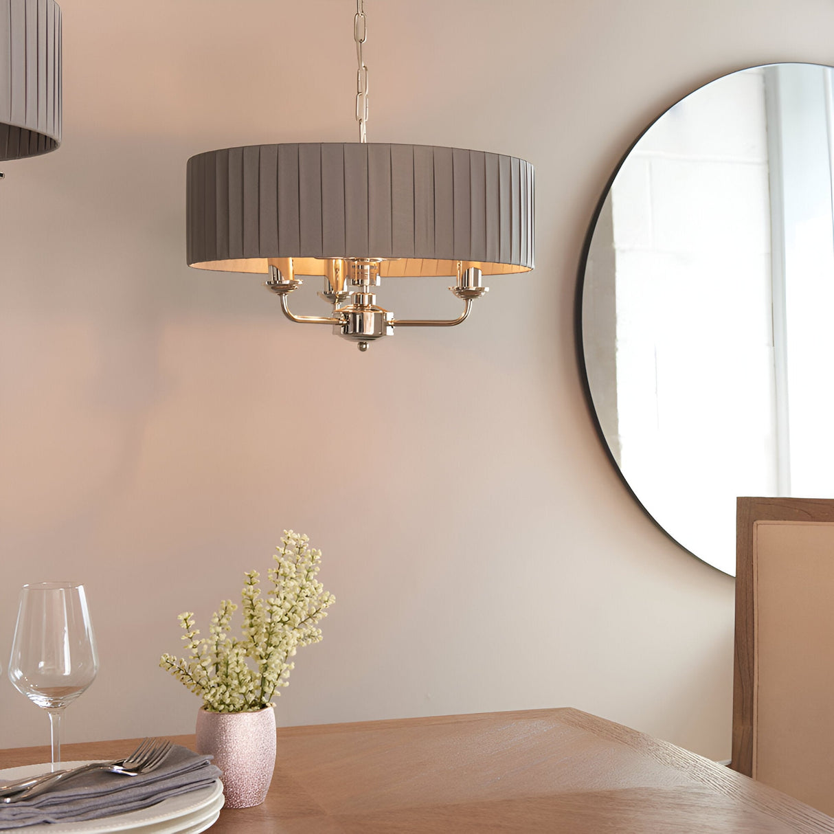 Brenley V2 3 Light Pendant in nickel with a pleated grey fabric drum shade, hung on a chain above a dining table near a round mirror.