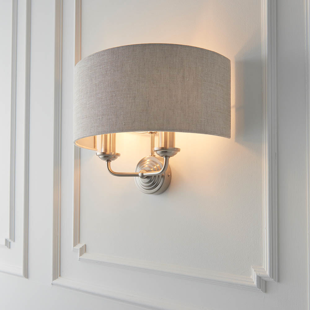 Brenley 2 Light Wall Light in chrome and brass with a curved natural linen shade over twin candle-style lamps, mounted on white panelled wall moulding.