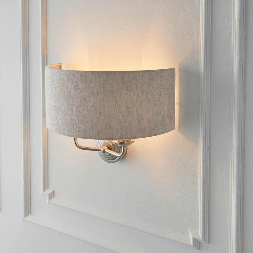 Brenley 2 Light Wall Light in chrome and brass with a curved natural linen shade, mounted on a white panelled wall showing a warm upward glow.