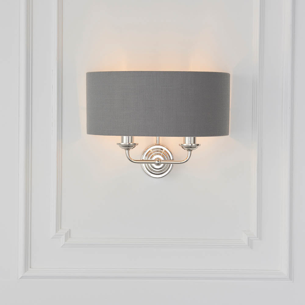 Brenley 2 Light Wall Light in nickel with a grey fabric drum shade, twin arms on a round backplate, mounted on a white panelled wall.