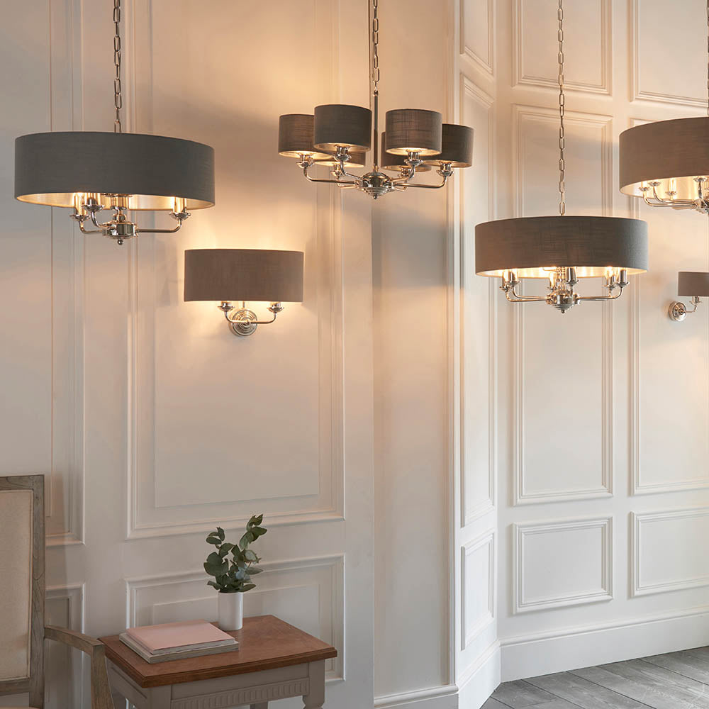 Brenley 2 Light Wall Light in nickel with a grey drum shade, shown mounted on a panelled wall beside matching pendant chandeliers.