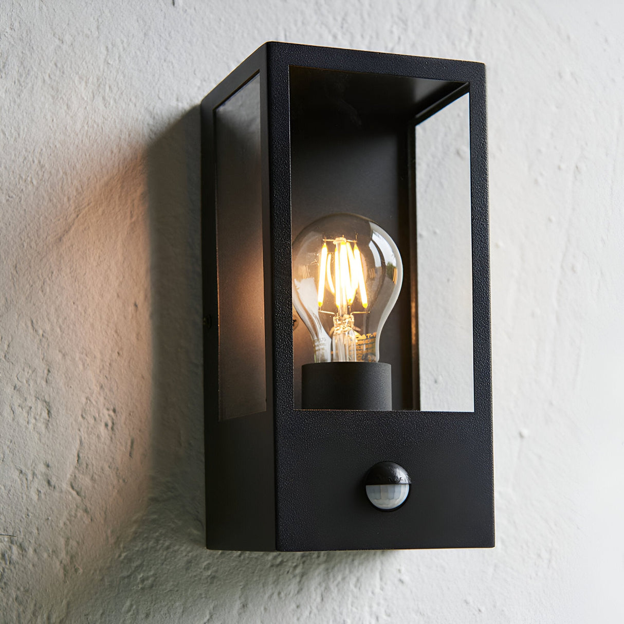 Castwood Outdoor Wall Light with PIR Sensor in matt black, box lantern with glass panels and lit filament bulb, mounted on a white textured wall.