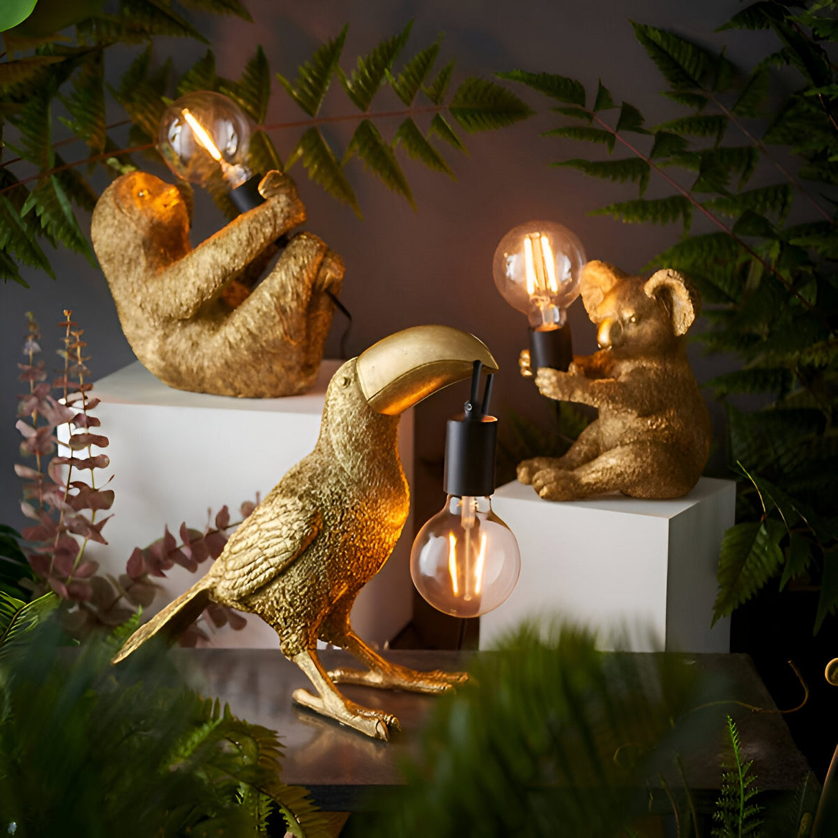 Cranmont Toucan Table Lamp in gold resin with matt black E27 holder and exposed filament bulb, shown among sloth and koala lamps in a foliage setting.