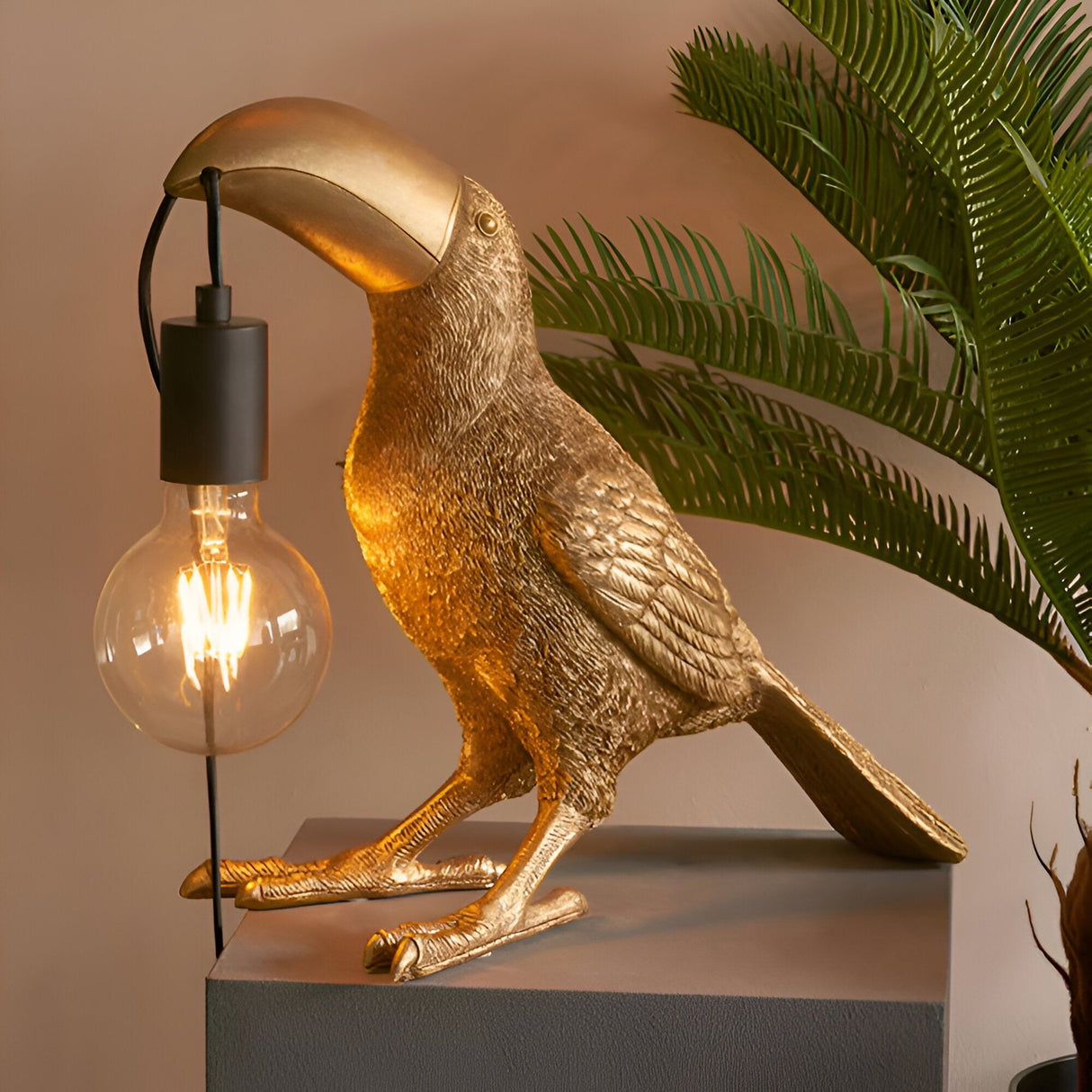 Cranmont Toucan Table Lamp in gold resin with matt black E27 bulb holder and exposed filament globe, shown on a plinth beside a palm plant.