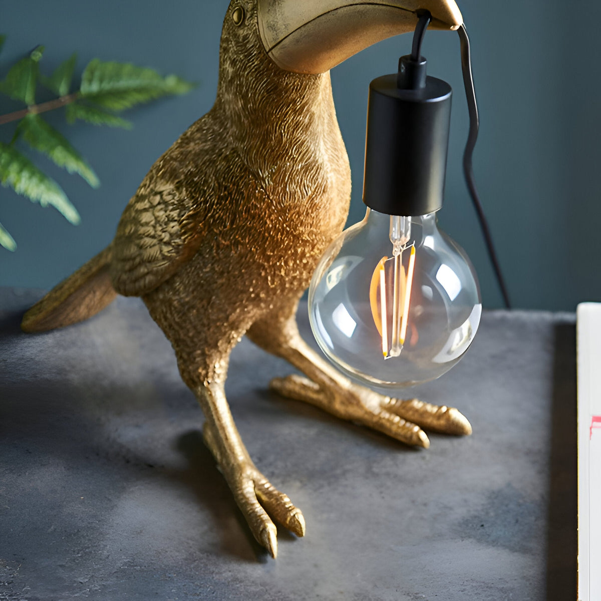 Cranmont Toucan Table Lamp in gold resin with black E27 bulb holder and exposed globe bulb, shown on a dark surface with fern leaves.