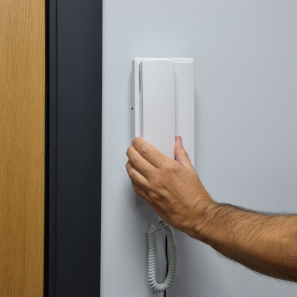Hand lifting the white wall-mounted handset of the AVSL Mercury 2-Way Intercom, with coiled cable hanging below beside a wooden door frame.
