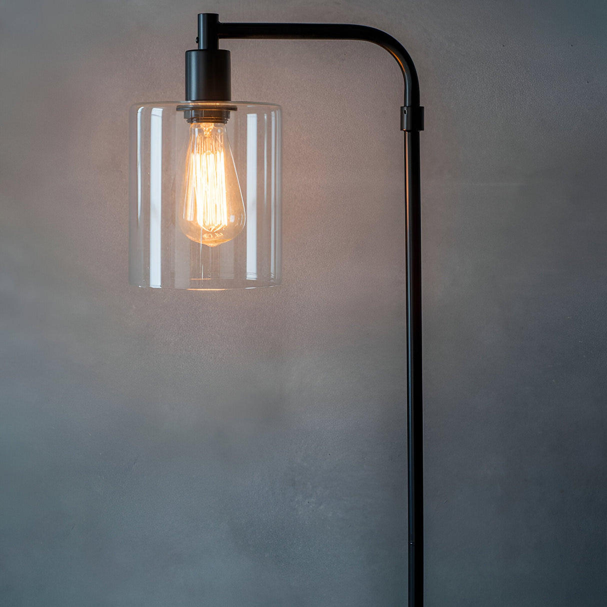 Cranmere Floor Lamp in matt black metal with a clear glass cylinder shade and glowing filament E27 bulb, shown against a plain grey background.