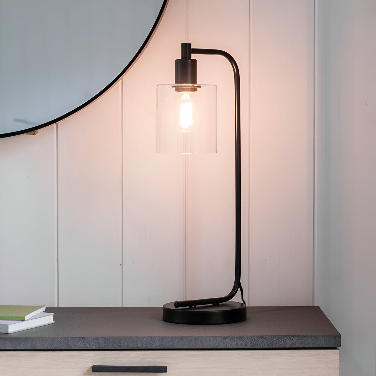 Endon Cranmere table lamp in matt black with clear glass cylinder shade and exposed filament bulb, on a dark surface beside a round mirror.