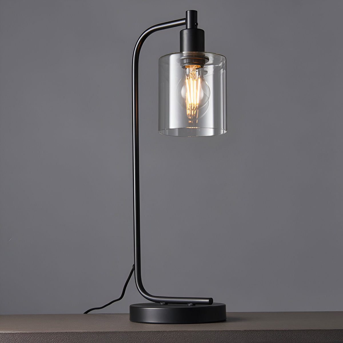 Endon Cranmere table lamp in matt black metal with an arched stand, clear glass cylinder shade and exposed filament E27 bulb, on a grey background.