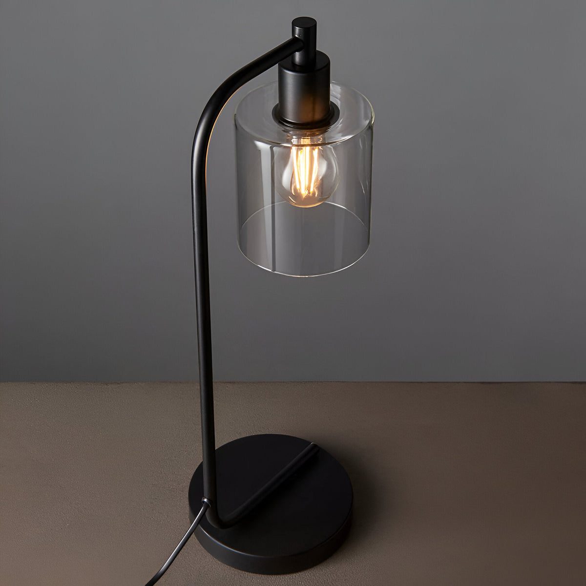Endon Cranmere table lamp in matt black with an arched stem, round base, clear glass cylinder shade and exposed filament bulb on a neutral surface.