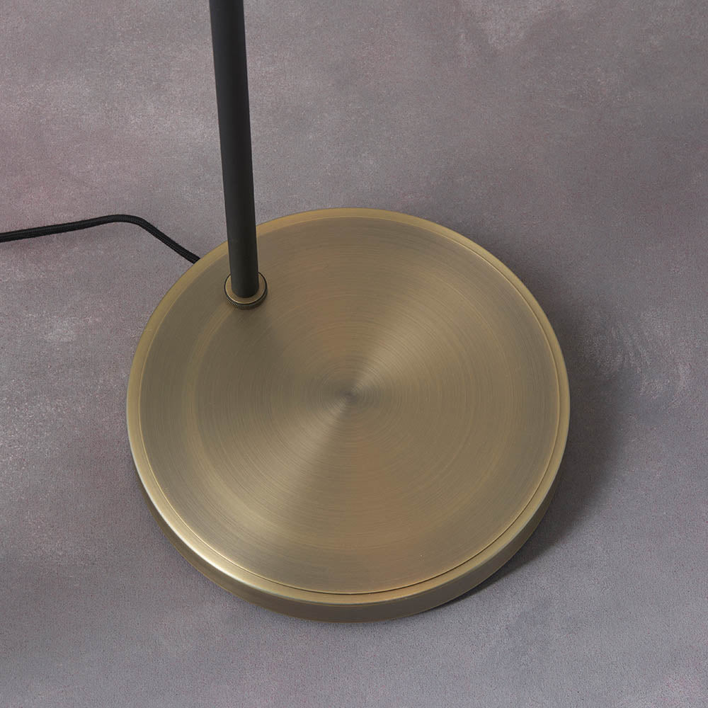 Close-up of the Carlbourne Floor Lamp's round brass base with black metal pole and cable, shown on a grey surface.
