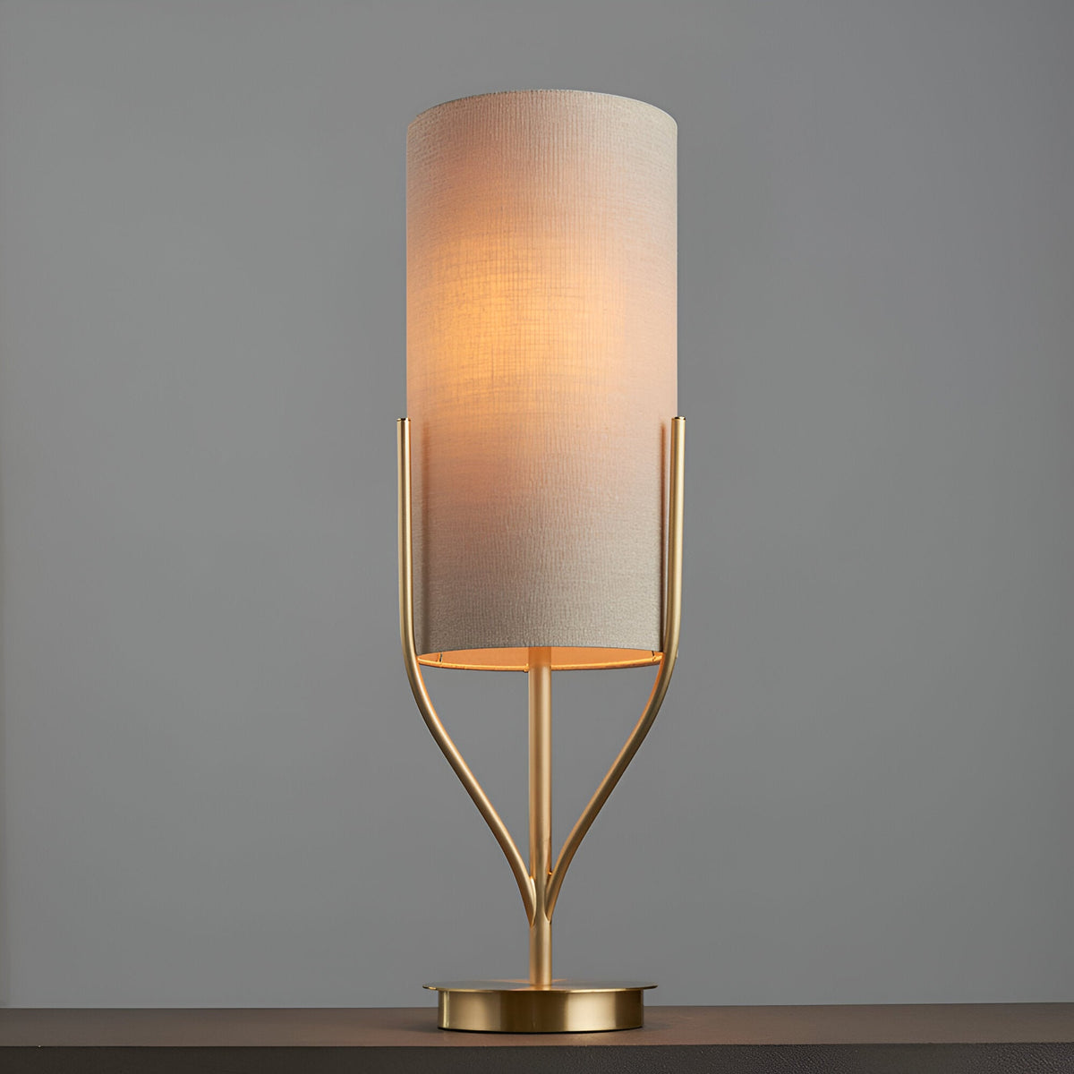 Endon Bramdon table lamp with satin brass forked base and tall natural linen cylinder shade, illuminated against a plain grey background.